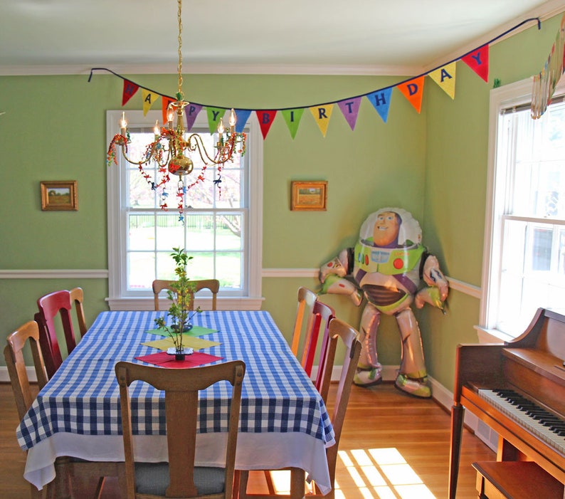 May include: A green and white checkered tablecloth covers a table set for a birthday party. A colorful banner with the words "Happy Birthday" hangs above the table. A large inflatable Buzz Lightyear toy stands in the corner of the room.