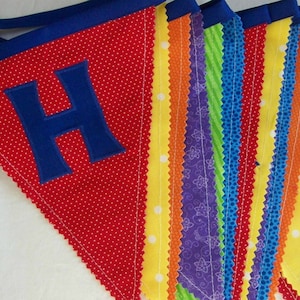 May include: A red fabric banner with white polka dots and a blue felt letter 'H'. The banner is made of multiple strips of fabric in various colors and patterns, including yellow, orange, purple, green, and blue.