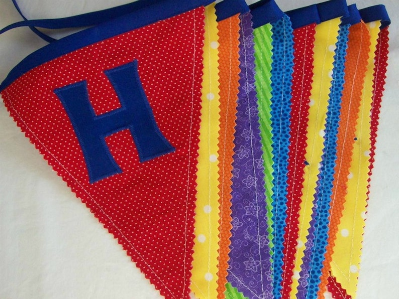 May include: A red fabric banner with white polka dots and a blue felt letter 'H'. The banner is made of multiple strips of fabric in various colors and patterns, including yellow, orange, purple, green, and blue.