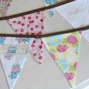 May include: A bunting banner with a brown ribbon and triangular flags made of different floral patterned fabrics. The flags are in shades of pink, blue, yellow, and white.