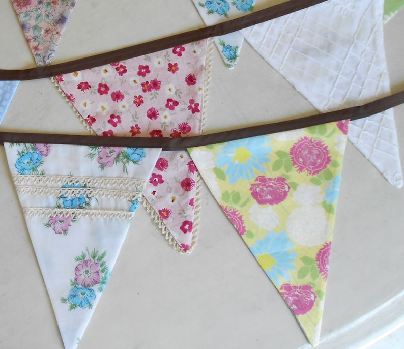 May include: A bunting banner with a brown ribbon and triangular flags made of different floral patterned fabrics. The flags are in shades of pink, blue, yellow, and white.