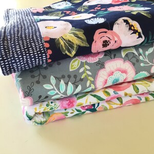 May include: A stack of folded fabric with floral patterns. The top layer features a navy blue background with pink and white flowers. Below are layers with gray, and white backgrounds, each with different floral designs. The top layer has a contrasting dotted pattern.