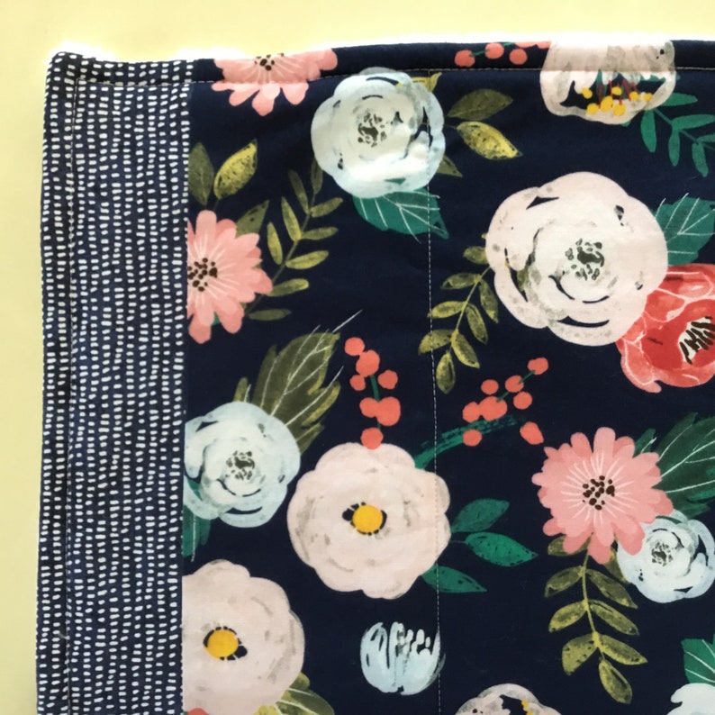 May include: Close-up of a floral fabric with a navy blue background. The fabric features large white, pink, and red flowers with green leaves. A border of navy blue fabric with white dots is on the left side.