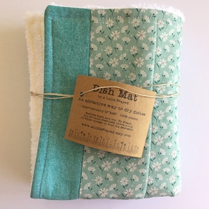 May include: A teal and white dish mat with a floral pattern. The mat is tied with twine and has a brown tag that reads "Dish Mat by a Little Frayed. An attractive way to dry dishes. Approximately 18"x22", 100% cotton. Machine wash and dry. No bleach, but for extra freshness, throw a cup of white vinegar in with the detergent. www.aLittleFrayed.etsy.com"