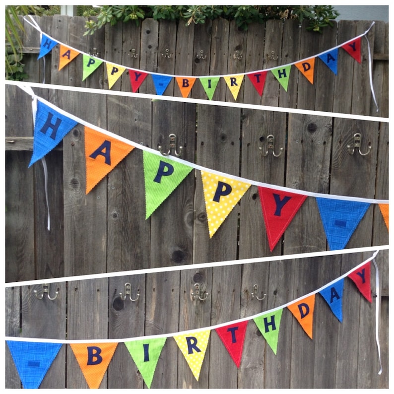 May include: A colorful fabric banner with the words "Happy Birthday" strung on a white ribbon. The banner is made of triangular flags with different colors and polka dots. The letters are black and the background is a wooden fence.