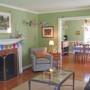 May include: A green living room decorated for a birthday party with a fireplace, a couch, a chair, a coffee table, and a dining room in the background. The fireplace has a brick surround and a brass fire screen. The couch is black leather and the chair is gray with a patterned cushion. The coffee table is glass with a metal frame. The dining room has a table with a checkered tablecloth and chairs. There is a banner hanging on the wall that says "LUKE".