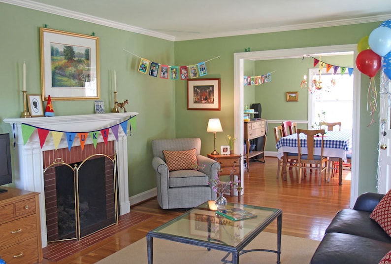 May include: A green living room decorated for a birthday party with a fireplace, a couch, a chair, a coffee table, and a dining room in the background. The fireplace has a brick surround and a brass fire screen. The couch is black leather and the chair is gray with a patterned cushion. The coffee table is glass with a metal frame. The dining room has a table with a checkered tablecloth and chairs. There is a banner hanging on the wall that says "LUKE".