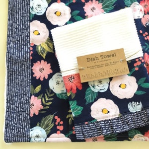 May include: A set of dish towels featuring a floral pattern with pink and white flowers on a navy blue background. One towel is white with a ribbed texture. A tag reads "Dish Towel" with care instructions.