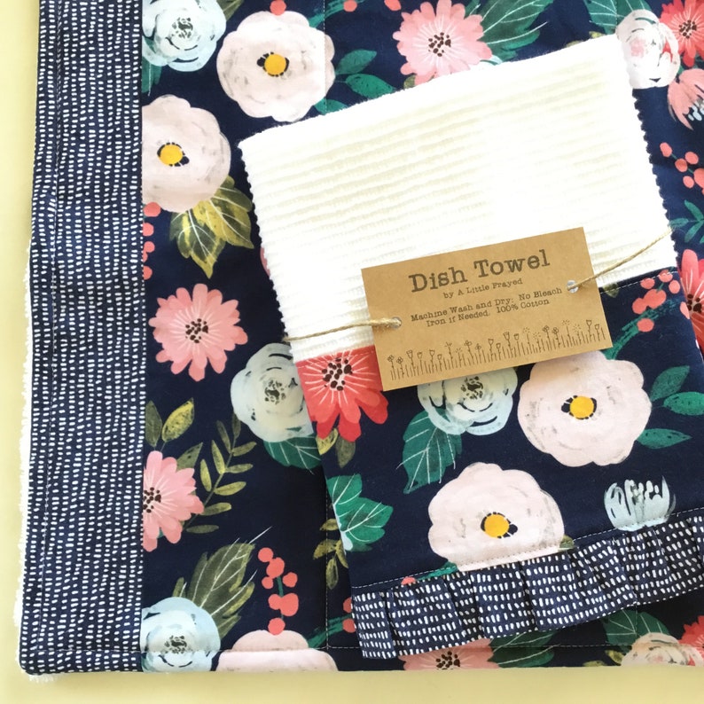 May include: A set of dish towels featuring a floral pattern with pink and white flowers on a navy blue background. One towel is white with a ribbed texture. A tag reads "Dish Towel" with care instructions.