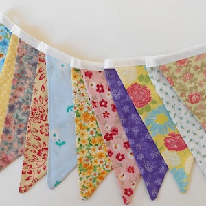 May include: A colorful fabric banner with a white trim. The banner is made of 10 triangular flags, each with a different floral pattern. The flags are sewn together with white thread. The banner is perfect for adding a touch of whimsy to any party or event.
