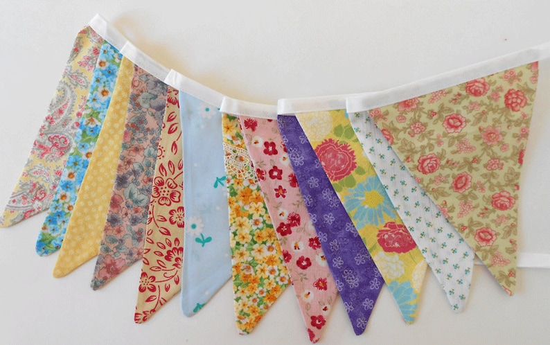 May include: A colorful fabric banner with a white trim. The banner is made of 10 triangular flags, each with a different floral pattern. The flags are sewn together with white thread. The banner is perfect for adding a touch of whimsy to any party or event.