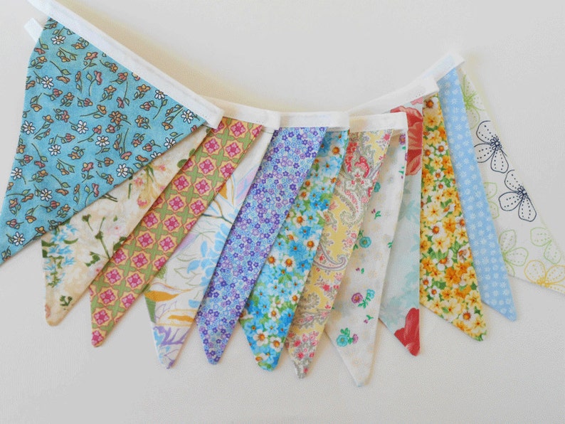May include: A bunting banner with 10 triangular flags. The flags are made of different fabrics with floral patterns in shades of blue, yellow, pink, and green.