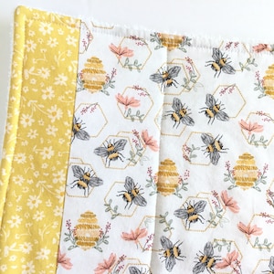 May include: A white fabric kitchen pot holder with a yellow border. The fabric has a repeating pattern of honeycombs, bees, and flowers.