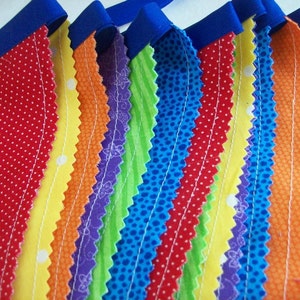 May include: A colorful fabric banner with alternating stripes of red, yellow, orange, purple, green, blue, and red. The fabric is sewn together with white thread and has a scalloped edge.