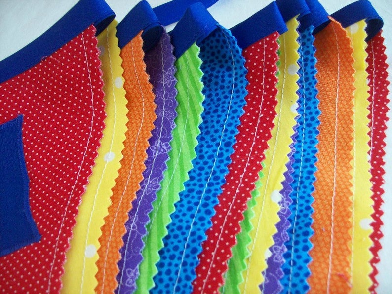 May include: A colorful fabric banner with alternating stripes of red, yellow, orange, purple, green, blue, and red. The fabric is sewn together with white thread and has a scalloped edge.