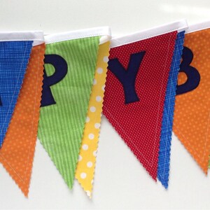 May include: A colorful fabric banner with the word "HAPPY" spelled out in blue felt letters. Each letter is on a separate triangular flag with a different patterned fabric. The flags are sewn together with white ribbon.
