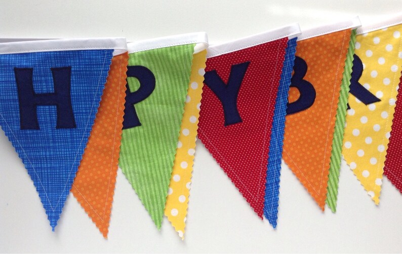 May include: A colorful fabric banner with the word "HAPPY" spelled out in blue felt letters. Each letter is on a separate triangular flag with a different patterned fabric. The flags are sewn together with white ribbon.