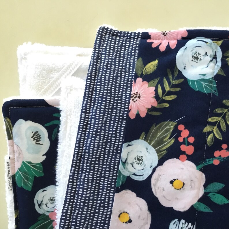 May include: Close-up of a floral patterned fabric with a navy blue background and white, pink, and green flowers. A white terry cloth is visible behind the fabric. The edge of the fabric has a dotted pattern.
