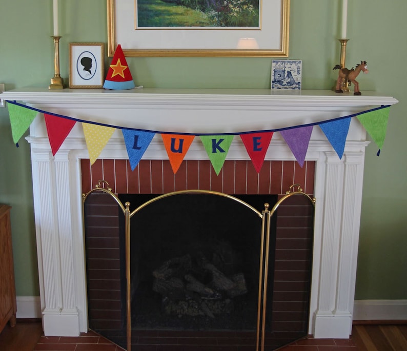 May include: A white fireplace mantel with a banner that reads "LUKE" in colorful letters. The banner is made of fabric and is strung with colorful yarn. There is a red and white star-shaped hat on the mantel, as well as a framed picture and a small wooden horse figurine.