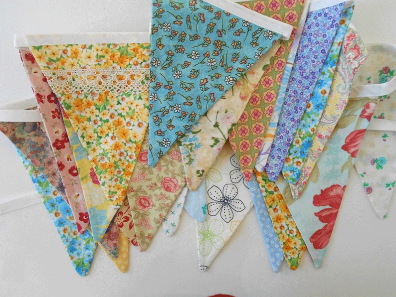 May include: A collection of colorful fabric bunting flags with floral patterns. The flags are sewn together with white ribbon and have a white lace trim.
