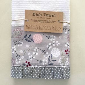 May include: A gray and white dish towel with a floral pattern and a ruffled edge. The towel has a brown tag that says "Dish Towel by A Little Frayed. Machine Wash and Dry: No Bleach. Iron if Needed. 100% Cotton."