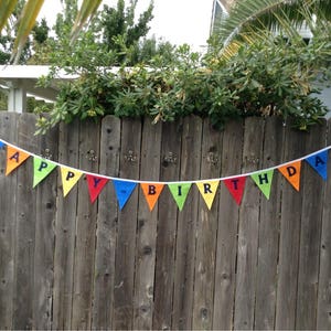 May include: A colorful Happy Birthday banner with rainbow-colored triangular flags strung together with white string. The banner is hanging on a wooden fence.