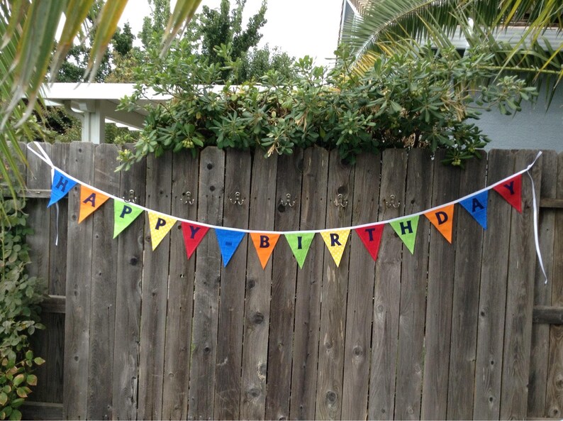 May include: A colorful Happy Birthday banner with rainbow-colored triangular flags strung together with white string. The banner is hanging on a wooden fence.