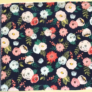 May include: A floral placemat featuring a navy blue background with a pattern of pink, white, and coral flowers, green leaves, and small red berries. The left side has a vertical stripe of white dots on a navy blue background. The placemat is rectangular.