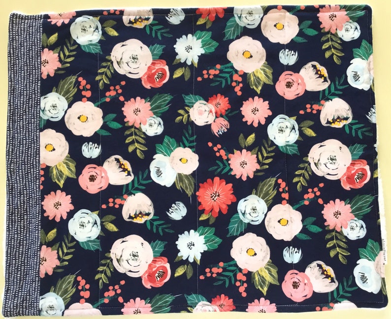 May include: A floral placemat featuring a navy blue background with a pattern of pink, white, and coral flowers, green leaves, and small red berries. The left side has a vertical stripe of white dots on a navy blue background. The placemat is rectangular.