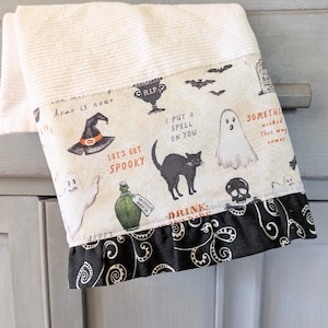 May include: A white Halloween-themed hand towel with a patterned border. The border features spooky illustrations of bats, ghosts, a black cat, and a witch's hat. Text includes "Let's get Spooky" and "Drink". A black decorative trim completes the design.