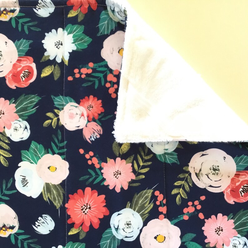 May include: A navy blue floral blanket with a pattern of pink, white, and red flowers and green leaves. The blanket has a soft, cream-colored, plush backing. The blanket is likely made of a soft, comfortable material.