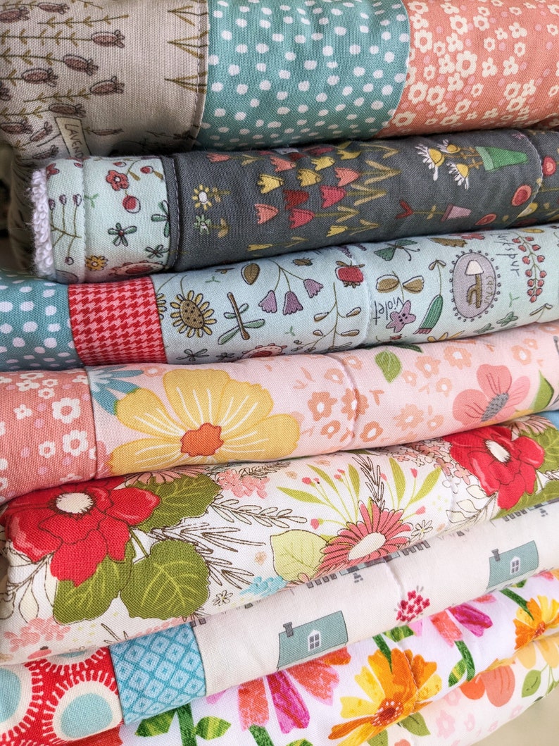 May include: A stack of folded, colorful fabric quilts. Each quilt features a patchwork design with floral and geometric patterns in shades of pink, green, yellow, and blue. The quilts are neatly stacked, showcasing the variety of designs.