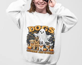 May include: White sweatshirt with a graphic featuring the text "DOGS because HUMANS ARE SPOOKY". The design includes a ghost and four dogs wearing orange bandanas and sitting in front of pumpkins. The person in the image is wearing a black beanie.