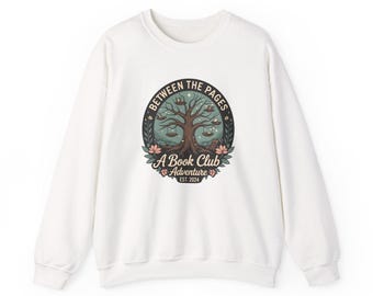 May include: White sweatshirt with a circular graphic featuring a tree with books and the text "BETWEEN THE PAGES A Book Club Adventure EST. 2024". The design includes floral and celestial elements.