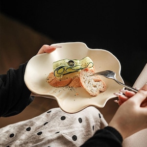Könnte beinhalten: Ein beigefarbener, organisch geformter Teller enthält eine kleine Vorspeise. Die Vorspeise besteht aus getoastetem Brot, belegt mit einer Gurkenrolle und einer kleinen schwarzen Olive. Eine Gabel ist bereit, einen Bissen zu nehmen. Der Teller wird von einer Person gehalten.