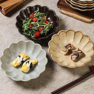 May include: A collection of ceramic serving dishes with a scalloped edge. One bowl holds a salad with tomatoes and blueberries. Two other bowls contain sushi. The dishes are in shades of brown, beige, and light blue.