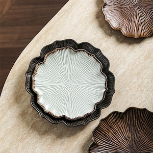 May include: A stack of three decorative ceramic plates with a scalloped edge. The top plate is white with a radial pattern, the middle plate is black with a brown rim, and the bottom plate is black. Two additional plates are visible.