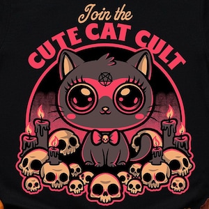 Puede incluir: Camiseta negra con un diseño de gato de dibujos animados y el texto "Join the Cute Cat Cult". El gato está rodeado de calaveras y velas, en tonos de negro, rosa y naranja. Un divertido gráfico temático de Halloween.