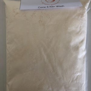 May include: A bag of Cone 6 Kiln Wash from Kinney Ceramic Supplies. The bag is filled with a light beige powder and is sealed in a clear plastic bag. The product label is white with black text and a logo.