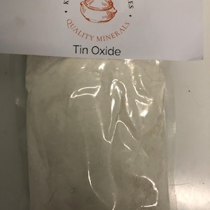May include: A clear plastic bag filled with a white powder, labeled "Tin Oxide." The bag is sealed and has a white paper label at the top with the text "Kinney Ceramic Supplies" and a drawing of a pot.
