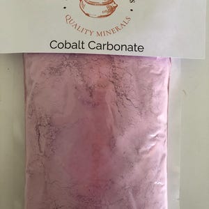 Puede incluir: Una bolsa de plástico transparente llena de polvo rosa, etiquetada como "Carbonato de Cobalto". La bolsa está sellada con una cabecera de papel blanco con el texto "Kinney Ceramic Supplies" y un dibujo de una olla.