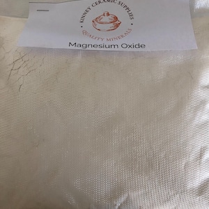 Pode incluir: Um saco branco de óxido de magnésio para materiais de cerâmica. O saco tem uma etiqueta de papel grampeada com o texto "Kinney Ceramic Supplies" e "Quality Minerals". A etiqueta também apresenta um desenho de um pote.