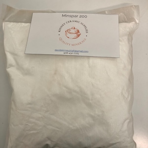 May include: A clear plastic bag filled with white powder, labeled "Minspar 200." A white card with the text "Kinney Ceramic Supplies" and a logo is attached to the bag. The bag is on a white surface.
