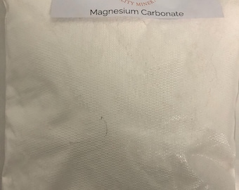 May include: A white bag of Magnesium Carbonate. The bag has a textured surface and a label at the top with the text "Kinney Ceramic Supplies Quality Minerals" and the product name.