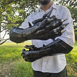 Peut inclure: Gantelets en métal noir avec des extensions pointues en forme de griffes. Les gantelets sont portés par une personne en chemise blanche et pantalon sombre. L'armure semble faite à la main et détaillée, avec un thème médiéval ou fantastique.