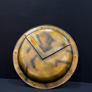 May include: A circular shield with a bronze finish, featuring a central V-shaped design. The shield has a weathered, antique appearance, with rivets around the edge. The design evokes a sense of history and ancient warfare.