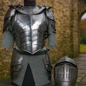 May include: A full suit of medieval armor, including a breastplate, chainmail, and helmet. The armor is made of dark metal and is displayed on a mannequin. The helmet has vertical slits for vision.