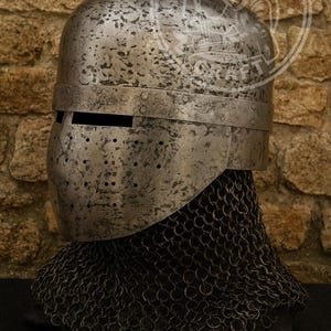 Puede incluir: Un casco metálico desgastado con visera y pequeños agujeros. El casco forma parte de una armadura, diseñado para proteger la cabeza. Se ve una cofia de cota de malla debajo del casco. El fondo es una pared de piedra.