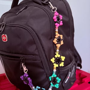 Puede incluir: Una mochila negra SwissGear con una larga cadena decorativa de cuentas unida al asa superior y a una cremallera lateral. La cadena consta de varios grupos en forma de flor hechos de cuentas negras, rosas, naranjas, amarillas, turquesas y azul claro.