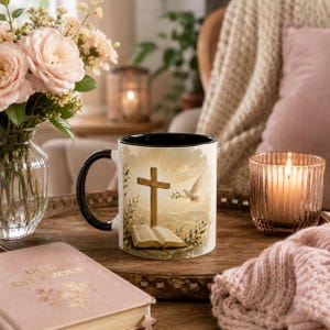 Peut inclure: Un mug en céramique avec une anse et un bord noirs, orné d'une illustration imprimée représentant une croix en bois, un livre ouvert et une colombe blanche sur un fond rayonnant et ensoleillé, avec des rayons dorés et des branches feuillues.